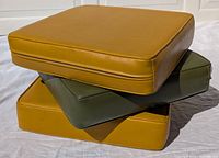 Three vinyl bench cushions stacked, two mustard yellow and one olive green, showing front and top surfaces with visible piping edges.