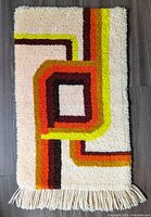 Full view of rectangular hooked textile with retro geometric design and fringed edge lying on dark floor.