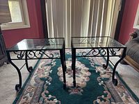 Pair of vintage side tables with glass tops and iron scrollwork legs photographed on a floral carpet.