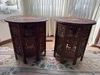 Front view of the pair of round ornately carved wood side tables placed on a floral pattern rug in front of window with sheer curtains.