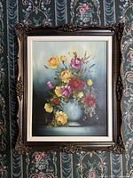Framed full view of floral still life oil painting in ornate frame against patterned wallpaper.