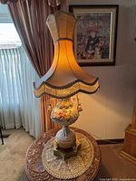 Full view of the lit lamp on round wooden table with lace doily, showing the fabric shade and ornate decorated base.
