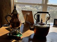 Full view of the copper coffee pot with burner and the copper tea pot on a wooden surface by a window, showing shape and size.