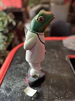 Side view of the green frog figurine dressed in white doctor coat with a stethoscope painted on, standing on a black base.