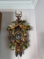 Full view of the Black Forest cuckoo clock showing detailed wood carvings, clock face with Roman numerals, and hanging pinecone weights.