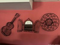 Full view of three metal wall decor pieces mounted on pink wall: guitar shape, decorative mirror with hinged panels, and sombrero.