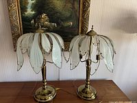 Pair of vintage floral table lamps with brass bases and glass petals shaped shades on a wooden surface with painting in background