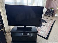 RCA 35 inch TV shown on a black TV stand with tinted glass door, located on carpeted floor with floral rug and wicker chair in background.