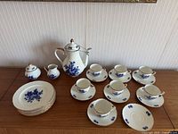 Full layout photo showing coffee pot, creamer, sugar bowl, cups, saucers and plates arranged on a wooden surface against a textured wall.