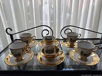 Five Tirschenreuth Bavaria espresso/tea cups with matching saucers, arranged on a black metal and glass table, showcasing the gold trim and scenic print on each cup.