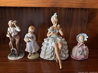 Various ceramic figurines on shelf including man with pitchfork, girl with basket, seated lady in ruffled dress with mirror, and girl in bonnet with flower basket.