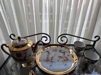 Full set of vintage Chinese porcelain teapot, plate, and two cups displayed on a metal tray in front of a sheer window curtain.