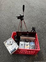Two camera tripods including a tall silver Lynx 1 model with black handle, and a smaller black tripod next to a red crate containing film and cameras.