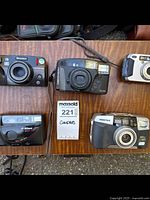 Four used 35mm film cameras including Olympus Multi AF, Nikon One Touch Discovery 290 Zoom, Pentax IQ Zoom 120mi, and Pentax IQ Zoom 95 WR placed on wood surface with a lot tag