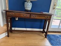 Front view of the wood console table against a blue wall and wooden floor, showing two drawers with brass pull handles.