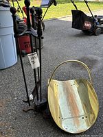 Set of four fireplace tools with decorative black handles and a brass-tone wood holder placed on pavement with garden equipment in background.
