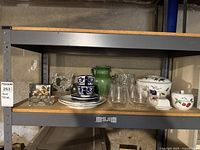 Shelf with assorted home decor items including crystal candlestick, gold votives, ceramic pitcher, plates, cups, and glasses