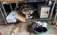 Wide angle showing Oxo mandoline slicer, chafing dish, spiralizer boxed, plastic bowl with coffee mugs, and tin pans on floor shelf