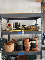 Metal shelving unit with assorted garden items on two shelves including terracotta and ceramic pots, sprinklers, and birdhouse mounting post.