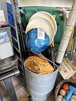 Photo showing a blue and black soccer ball, baseball gloves placed inside a metal bucket, and white frisbees leaning against a green folding chair.