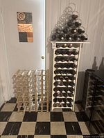 Photo of three wine racks side by side against a white door and wall: a black triangular plastic rack on top of a white plastic stacked rack loaded with wine bottles, and a wooden lattice rack next to them.