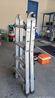 Folded aluminum multi-function ladder with articulating hinges, standing upright packed tightly in a garage.