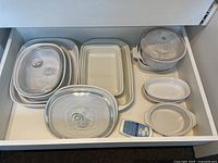 Top-down view of porcelain bakeware set in drawer, including large and smaller rectangular baking dishes, glass lids, white casserole pot with lid and side handles, and two small oval dishes.