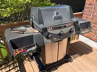 Front view of Broil King propane BBQ grill with closed lid showing three control knobs, side shelves, base cabinet with doors, set on deck with brick wall background.