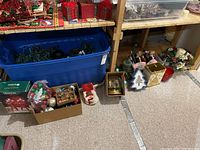 Wide view showing a large blue bin filled with Christmas lights, boxes of ornaments, snow globe, and other holiday décor items on the floor under wooden shelving.