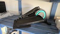 Side view of black and turquoise Cubii elliptical under desk pedal exerciser on blue surface.