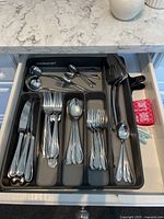 Fully opened drawer showing organized stainless steel cutlery including knives, forks, spoons, small spoons, and serving utensils in a grey plastic tray.