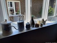 View of all seven ceramic decorative items arranged on a windowsill including watering can, lidded jar, candle, vase, cups, and bird figurine.