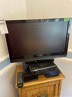 Front view of Dynex 22-inch TV on wooden surface with stand, remote placed nearby. Screen blank, frame glossy black with Dynex branding on bottom bezel.