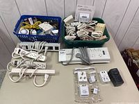 Lot overview showing two bins filled with assorted extension cords, power strips, and adapters along with several power strips and adapters laid out on a table.