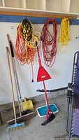Wide view showing extension cords hanging, brooms, dry mop, dustpans lined up against the wall in garage.