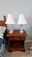Two clear glass base table lamps on a wooden nightstand next to a bed, each with white fabric shades.