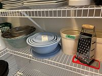 Shelf with multiple mixing bowls including gray metal and nested light blue bowls, two cream-colored pots, and metal grater with wooden handle and red base.