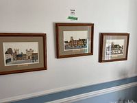 Three framed British needlepoint scenes hung on a white wall, showing castle, church, and village scenes with beige mats and wooden frames.
