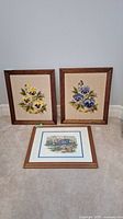 View of the three framed textile artworks: two with pansy embroidery and one with garden needlepoint in wooden frames.