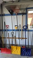 Photo of assorted outdoor tools hanging on wall including rakes, shovels, and snow shovels.