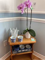 Orchid plant with pink flowers next to white ruffled glass vase and two small ceramic pots on wooden stand