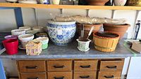 Shelves and table with collection of various planters including ceramic, terracotta, plastic and novelty shaped pots, gardening tools and burlap wraps on top shelf