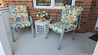 Two metal framed chairs with floral cushions, white woven round side table topped with a faux potted plant against brick wall on porch