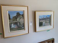 Two framed landscape paintings hanging on a wall with gold frames and white mats, featuring outdoor scenes with people and houses.