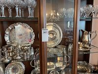 Wide view of cabinet shelf with various silver-plate items including trays, covered dish, candlesticks, salt and pepper shakers, and pitcher.