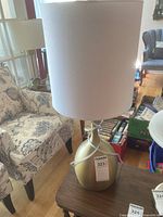 Full view of the table lamp showing the round brushed gold metal base and large white drum shade on a wooden side table with a floral upholstered chair in the background.