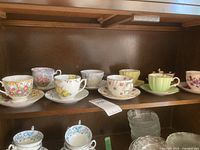 Multiple assorted vintage teacups with matching saucers displayed on a shelf, showing floral designs and various colors.