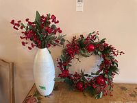 Front view of the white ceramic vase filled with red faux flowers and displayed with the decorative wreath featuring red berries and pine cones.