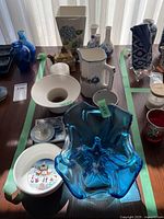 Photo showing an assortment of vases, a white bowl, porcelain teapot, small ceramic figurines, and glass items all laid out on a table.