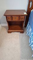 Front view of one wooden night table showing top surface, single drawer with two small brass-tone knobs and central handle, and lower open shelf. Medium brown finish and carpet floor.